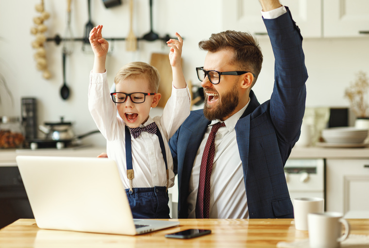 Symbolbild Freude im Homeoffice. Vater und Sohn im Business-Outfit mit Anzug, Krawatte und Nerd-Brille jubelnd vor dem Laptop zu Hause.